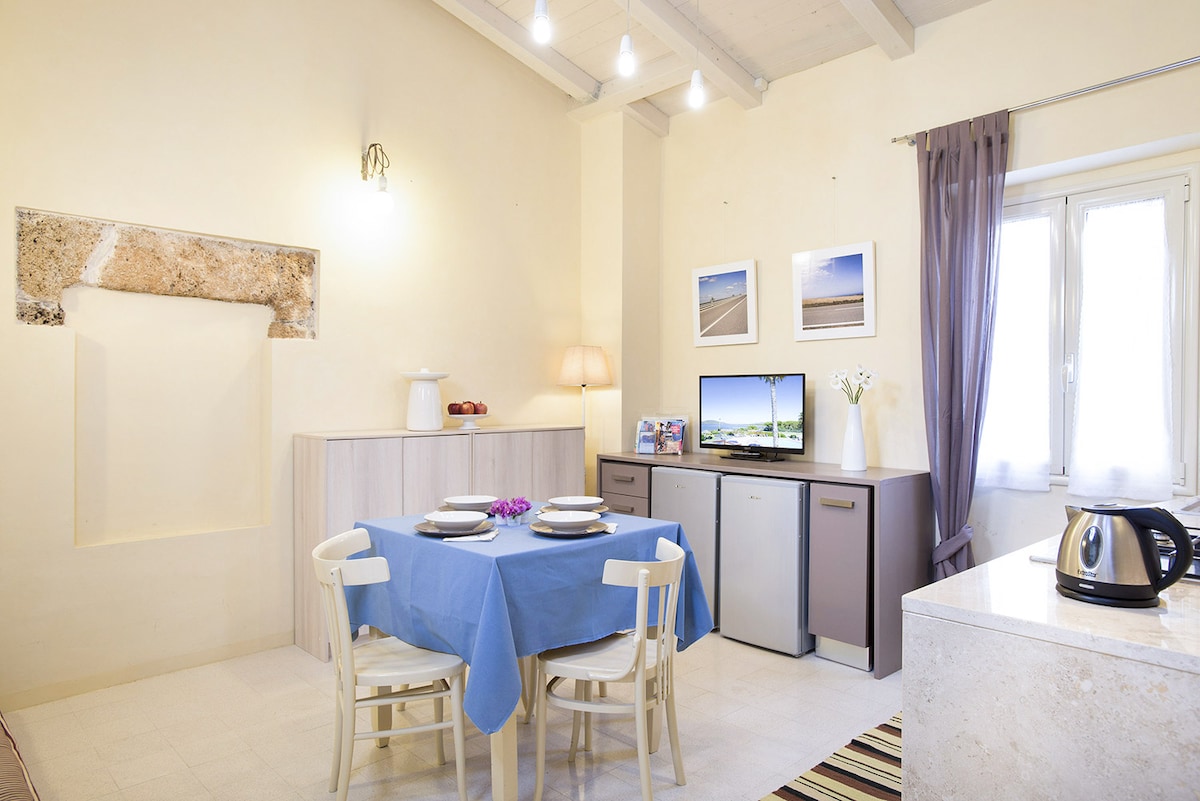 The dining area features a blue tablecloth covering a table set for four, surrounded by light wooden chairs. A sleek countertop is visible on the right, with modern appliances such as a kettle and a small refrigerator. Natural light enters through the window, brightening the room.