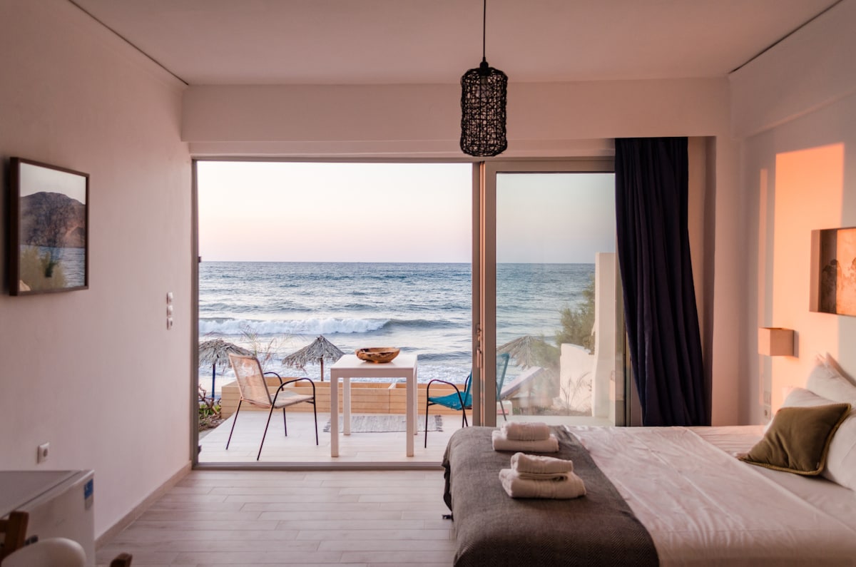 A serene sleeping area features a spacious bed with neatly arranged linens. Large glass doors offer an unobstructed view of the sea, with a small outdoor patio set that includes a table and chairs. Soft colors are complemented by natural light streaming in.