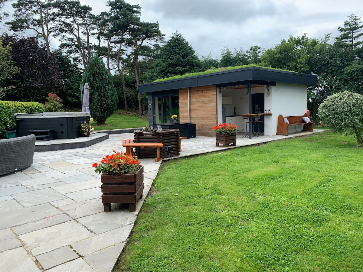 The image portrays a modern guest suite with a green roof, surrounded by well-maintained gardens. A stone patio features seating areas and flower beds, enhancing the outdoor space. A hot tub and a kitchenette area can be seen, providing guests with amenities for relaxation and entertainment.