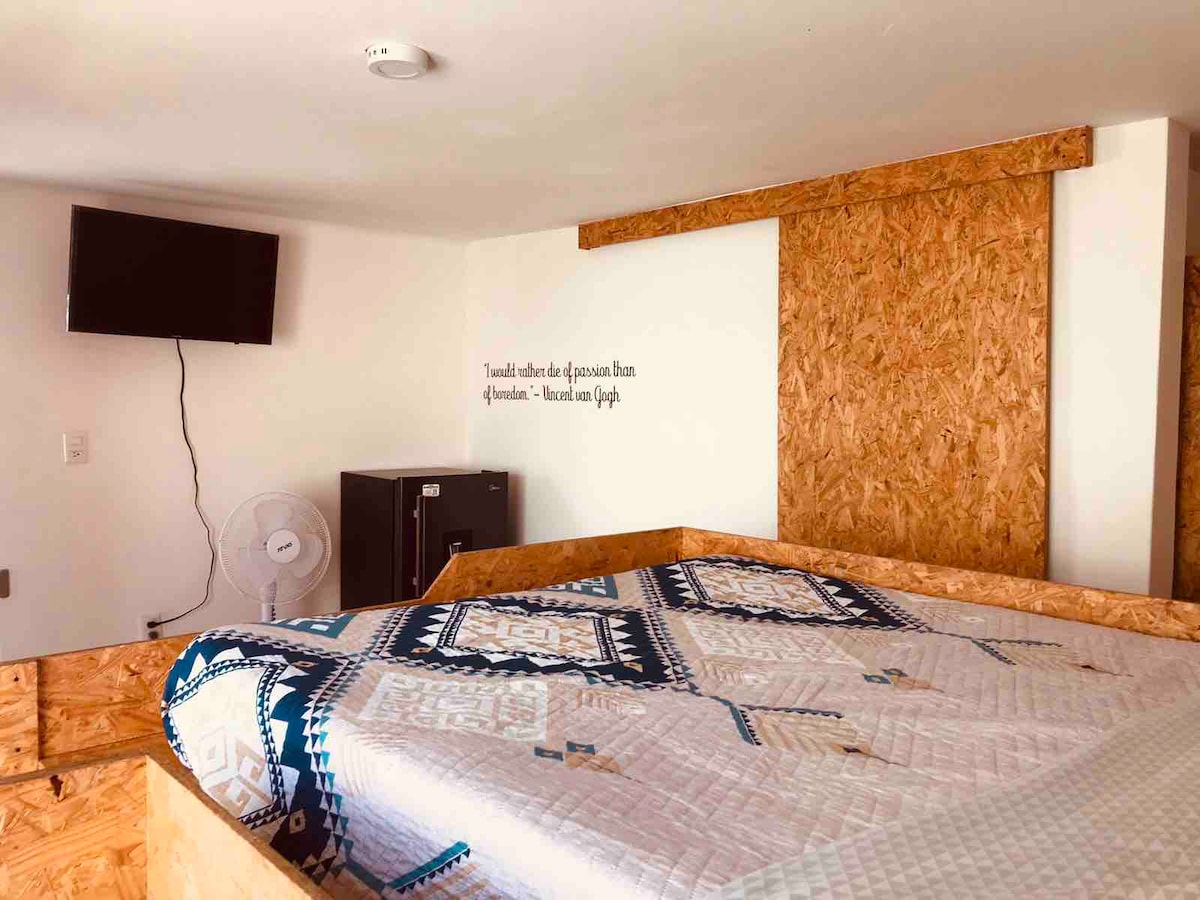 A cozy loft area is featured, showcasing a spacious bed with a patterned quilt. A flat-screen television is mounted on the wall, and a small fridge is positioned nearby. A wall fan provides additional comfort, while natural light enters through nearby windows.
