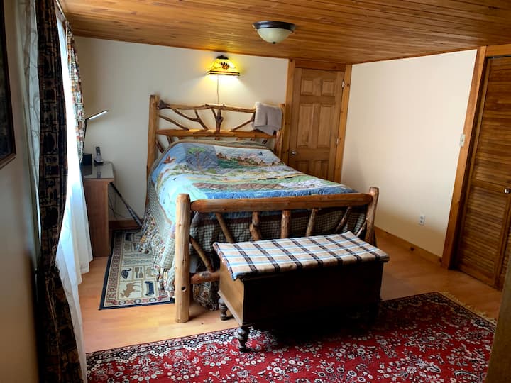 Spálňa inšpirovaná prírodou s manželskou posteľou, útulnou prikrývkou a veľkým oknom, ktoré prepúšťa prirodzené svetlo. Užite si pokojnú atmosféru, komodu na odkladanie a televízor na zábavu, vďaka čomu je tento priestor ideálny na oddych.