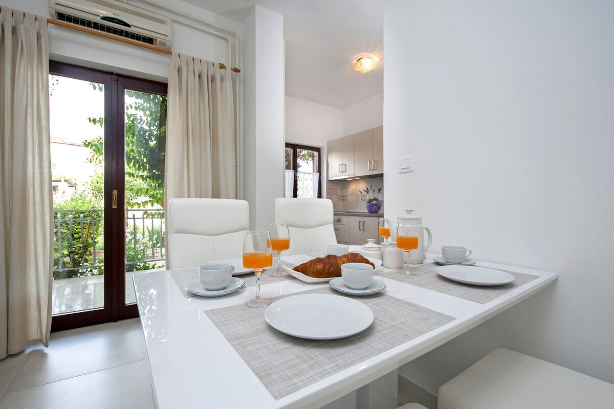 A bright dining area features a white table set with dishes, glasses, and a croissant. Sunlight filters through large windows, illuminating the space. Soft curtains frame the view of an outside balcony connecting to a lush garden.