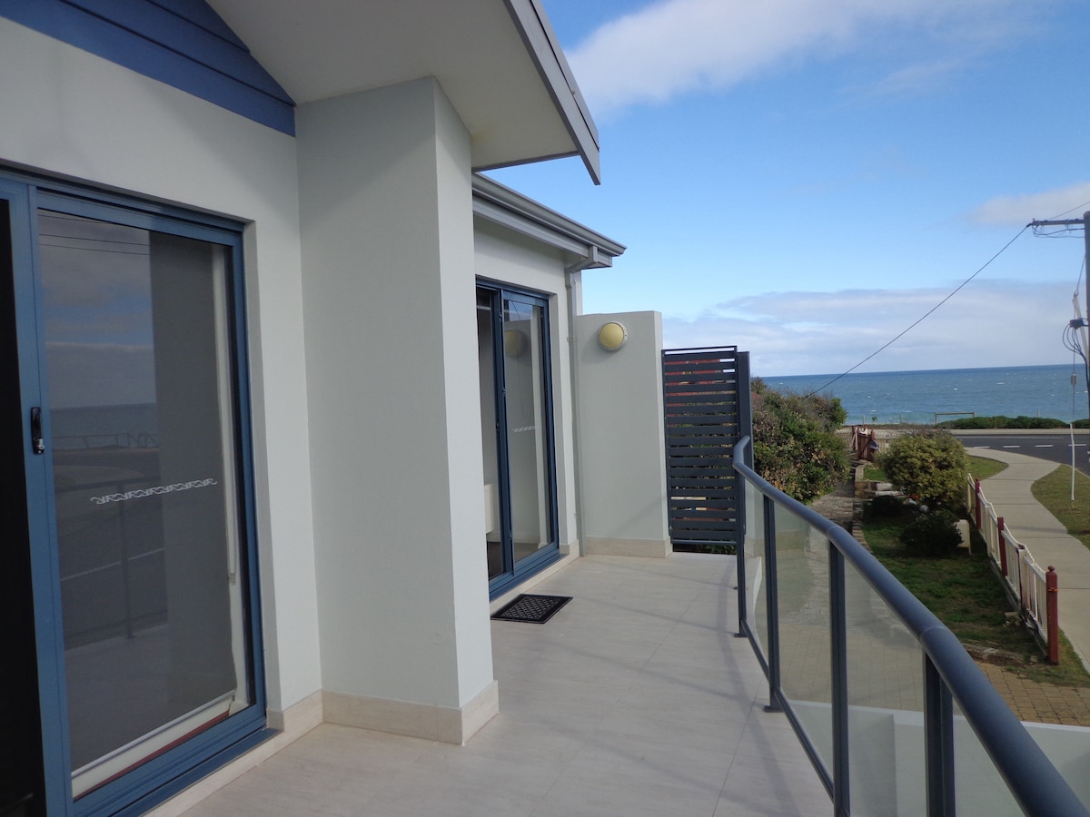 A spacious balcony is shown, featuring a glass railing that provides a clear view of the ocean. The exterior walls are painted in soft, neutral tones, complemented by large sliding glass doors that lead inside. The surroundings include green shrubbery and a nearby pathway.