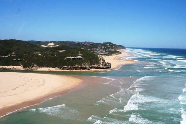 A panoramic view captures the coastline where sandy beaches meet the ocean waves. Gentle currents flow into a lagoon, bordered by lush greenery and residential areas in the distance. The clear blue sky complements the tranquil scene, bringing out the vibrant colors of the landscape.
