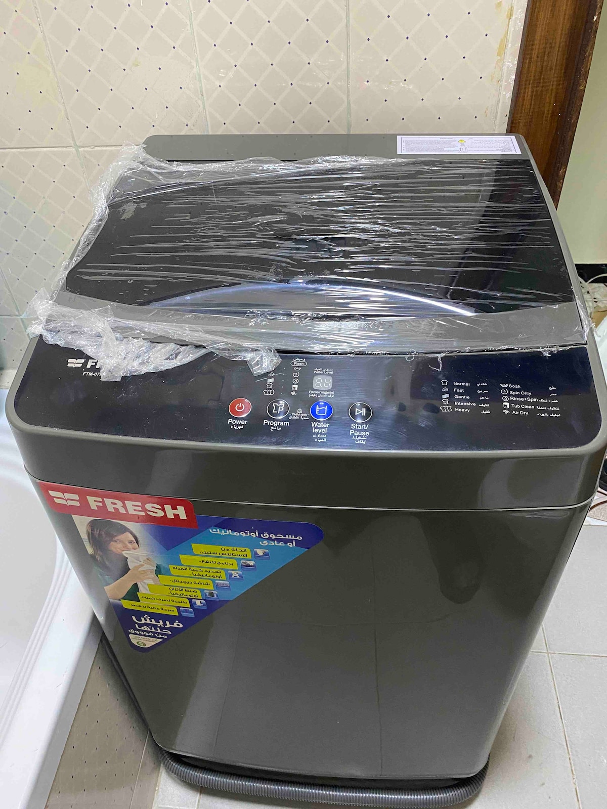 A modern washing machine is placed in a well-lit laundry area. The machine features a sleek dark exterior with a control panel displayed on the top, indicating various settings and functions. The surrounding tiles are neutral in color, enhancing the clean environment.