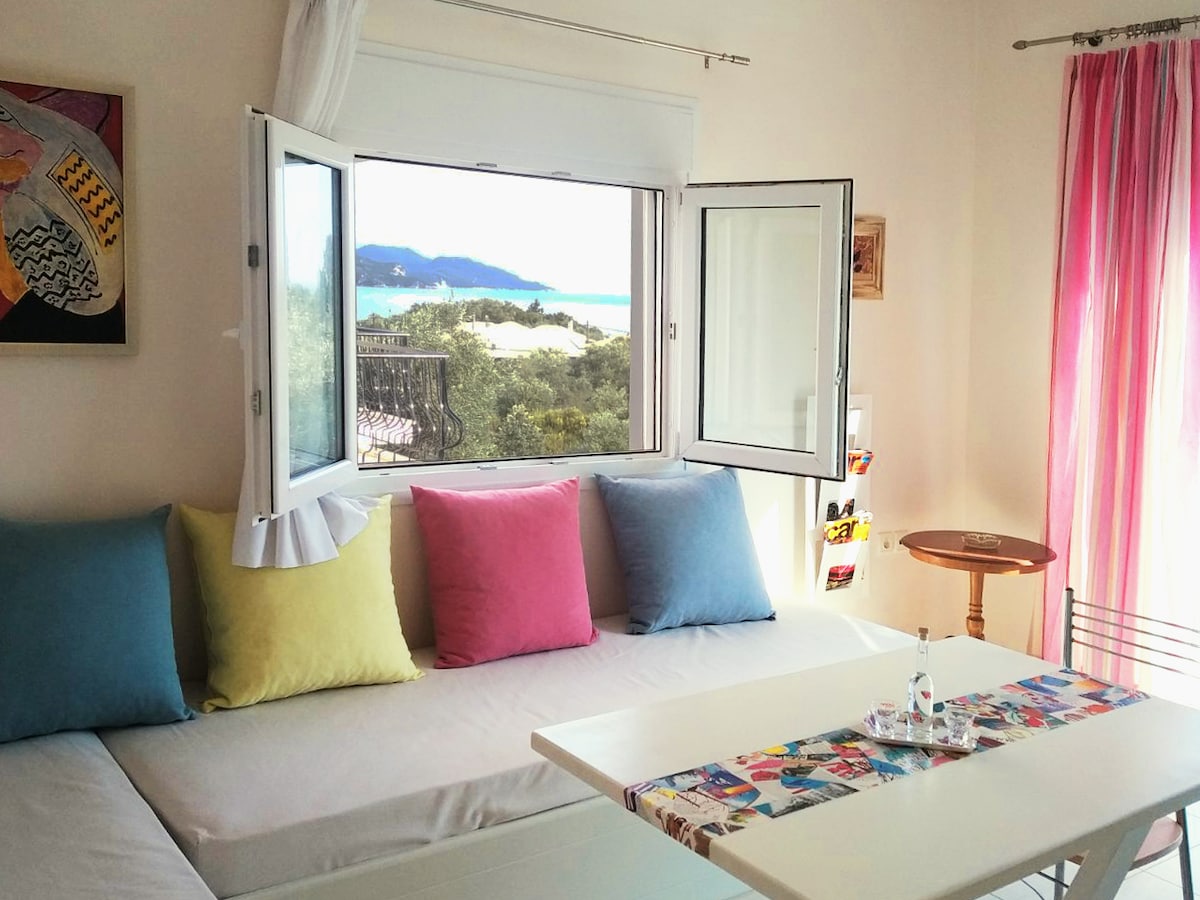 A light-filled seating area features a large window open to a scenic sea view. The window is framed by soft curtains. A white table, adorned with colorful artwork, is complemented by a cozy couch with pastel-colored cushions, creating a relaxed environment.