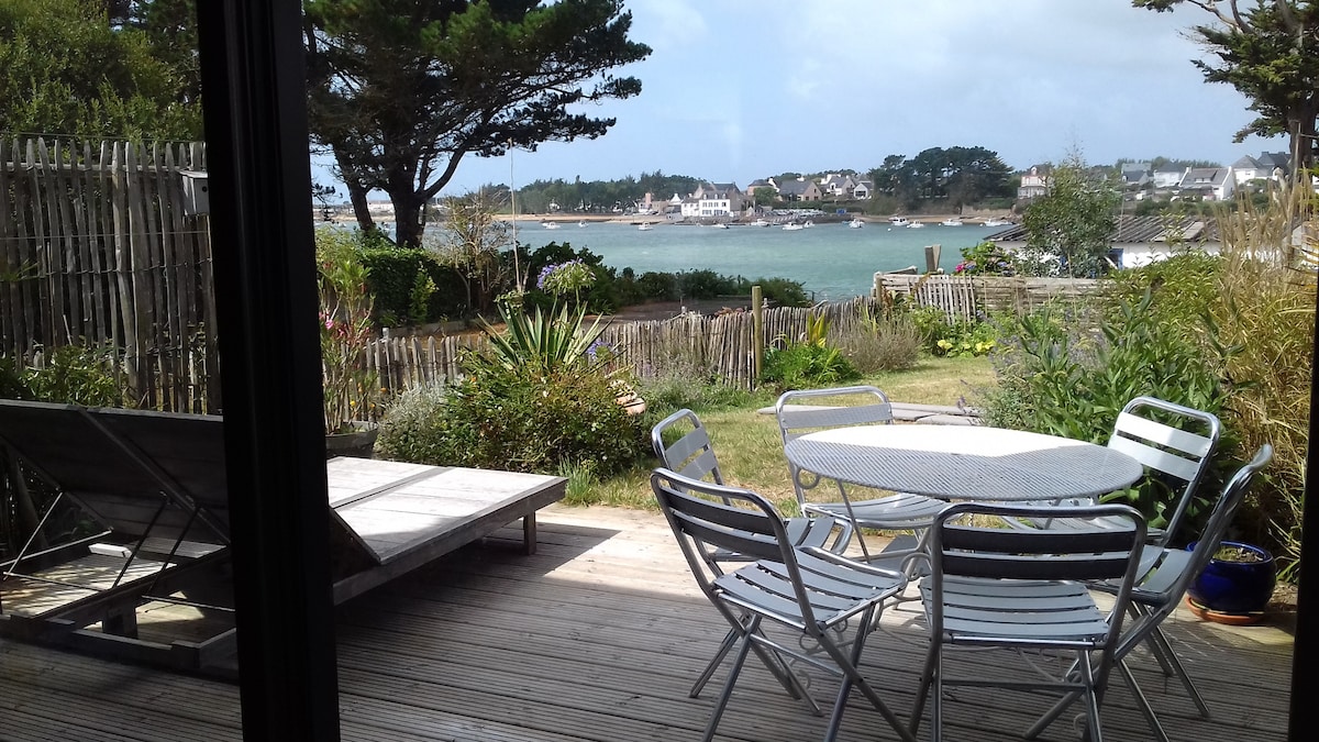A deck area features a round table surrounded by six sleek chairs, perfect for outdoor dining. A sun lounger rests nearby, while greenery frames the scene, leading to a view of the water and distant houses under a partly cloudy sky.