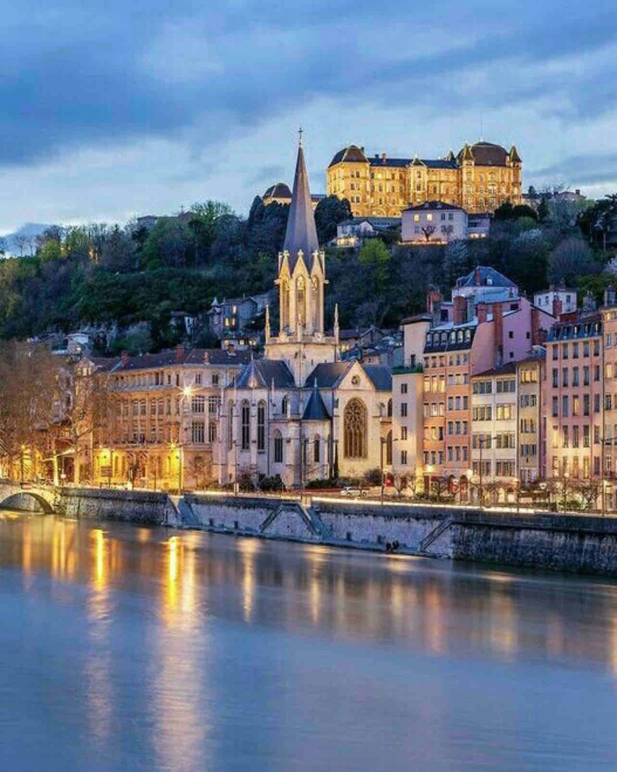 A riverside view showcases illuminated buildings along the water's edge, with a prominent church featuring a tall spire. Lush greenery is visible on the hillside behind, while the sky transitions from daylight to dusk, reflecting soft colors on the water's surface.