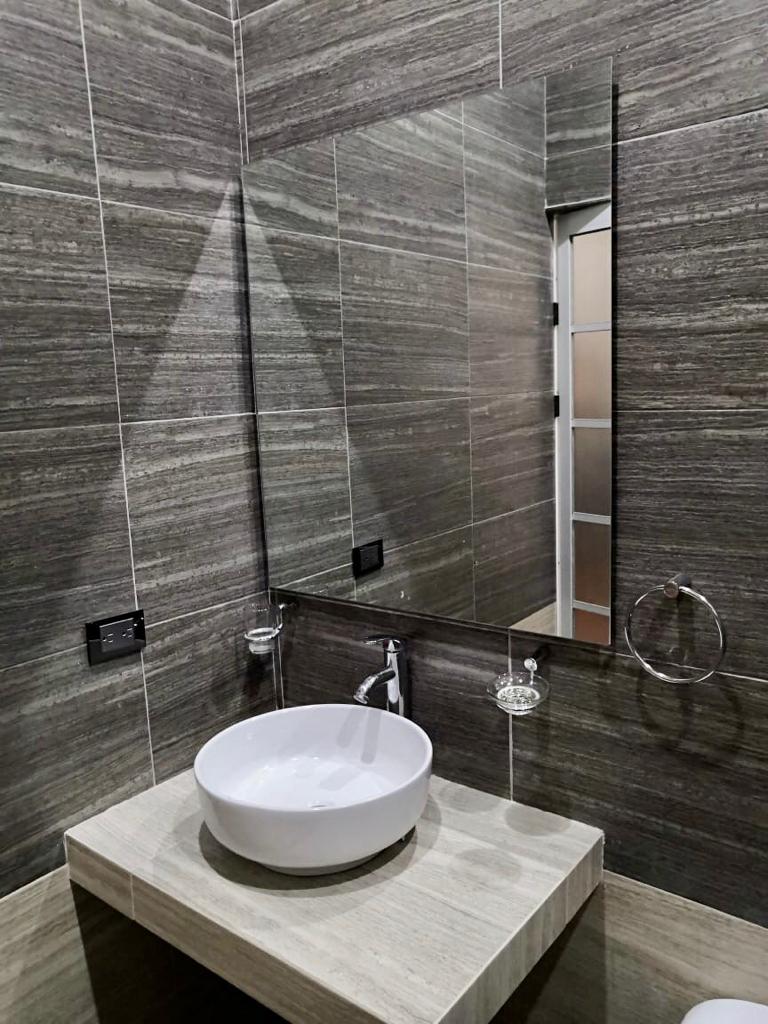 A modern bathroom features textured gray wall tiles, creating a sleek backdrop. A circular white sink is centered on a stone countertop, accompanied by a rectangular mirror above. A towel holder and faucet are positioned nearby, ensuring functionality and simplicity.