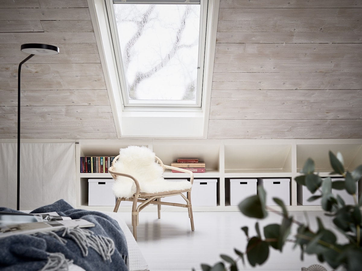 A cozy corner features a rattan chair adorned with a soft, white throw. Above, a skylight allows natural light to illuminate the space, highlighting the light wood paneling and simple bookshelves filled with books and storage boxes.