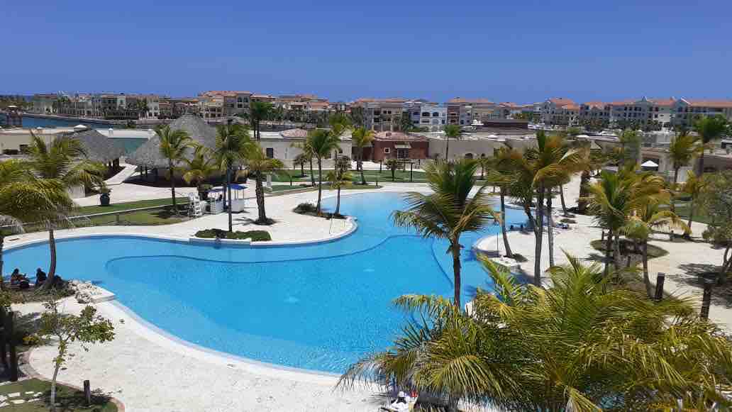 A spacious outdoor area is highlighted by a large swimming pool, surrounded by lush palm trees. Comfortable seating arrangements and sun loungers are positioned on the surrounding deck. Sunlight reflects off the water, creating a calming ambiance, while nearby buildings create a picturesque backdrop.