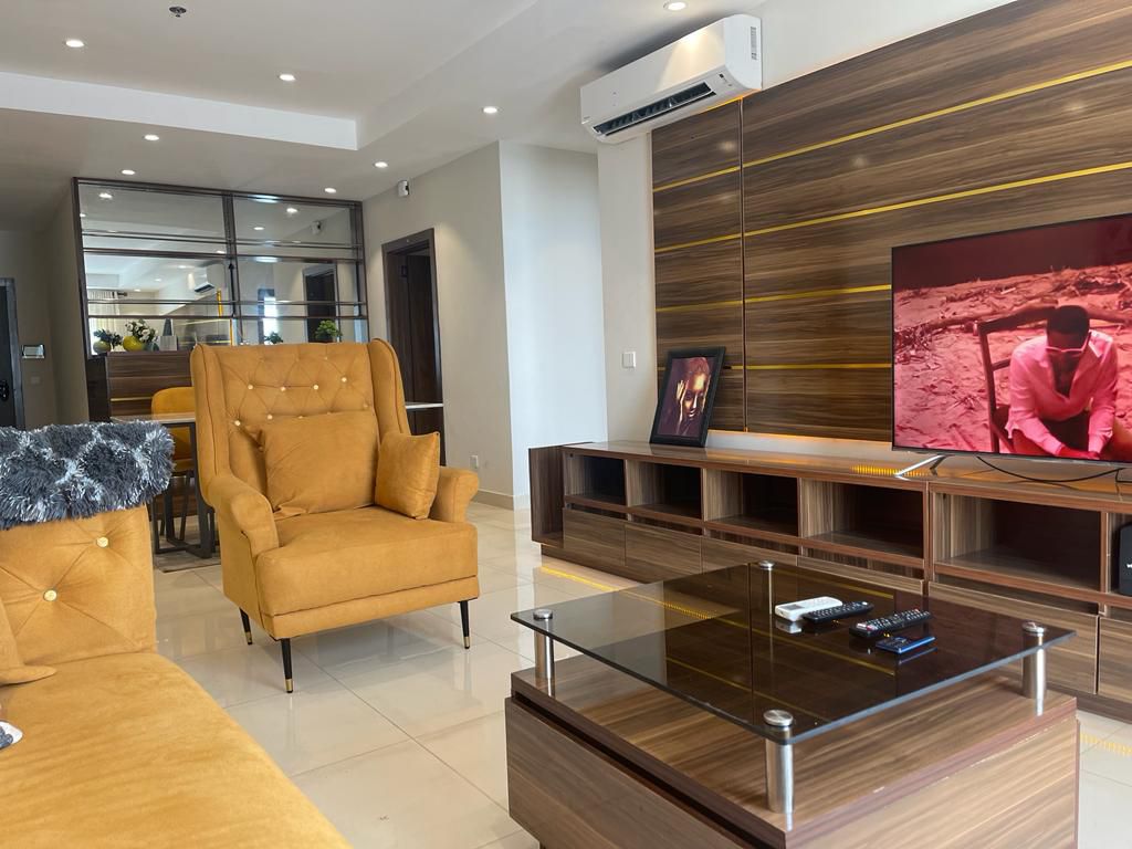 A modern living area is presented with a vibrant yellow armchair and a spacious sectional sofa adorned with a textured throw. A glass coffee table sits in front, accompanied by a media console that features a television displaying a scene in a warm colour palette.