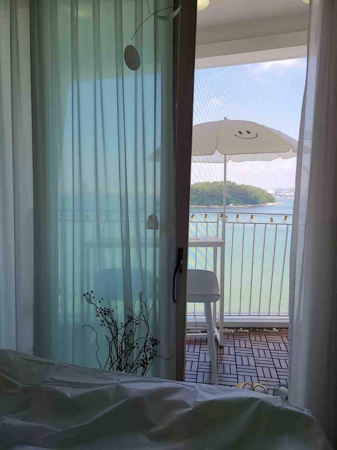A serene view is captured through sheer curtains, revealing a small balcony with a white chair and an umbrella. The scene overlooks tranquil waters and a distant island, creating a soothing backdrop from the indoor space.
