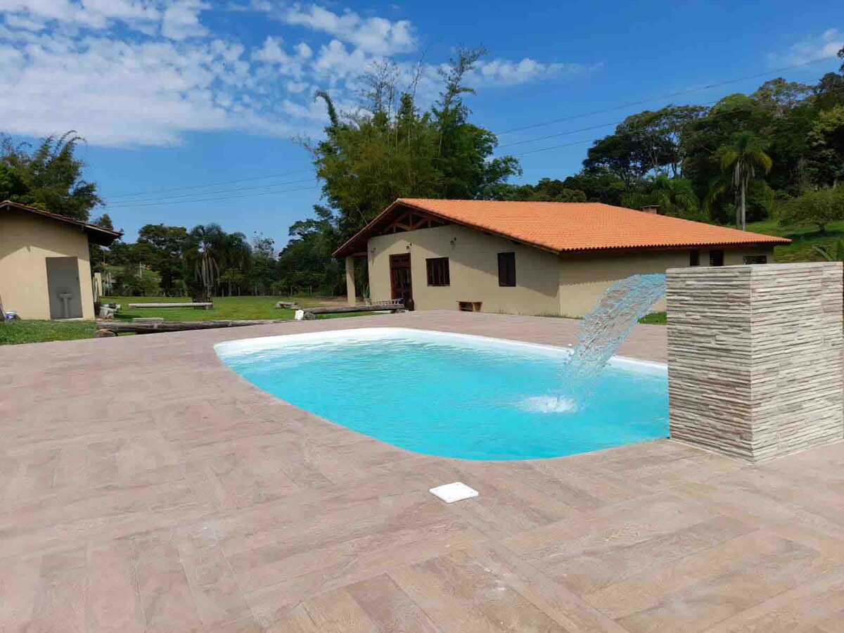 A refreshing swimming pool is highlighted, featuring a fountain that adds gentle movement to the clear water. The poolside is surrounded by a tiled surface, with a charming house visible in the background, set against a backdrop of lush greenery and a bright blue sky.