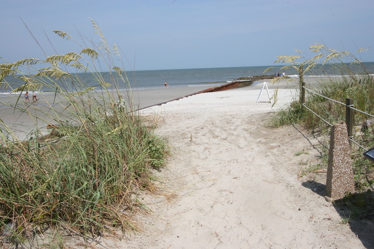 A sandy pathway leads through coastal grass toward the beach, with gentle waves visible in the distance. Sunlight illuminates the shoreline, where people can be spotted enjoying the water. The scene captures a serene coastal atmosphere, inviting exploration of the beach.