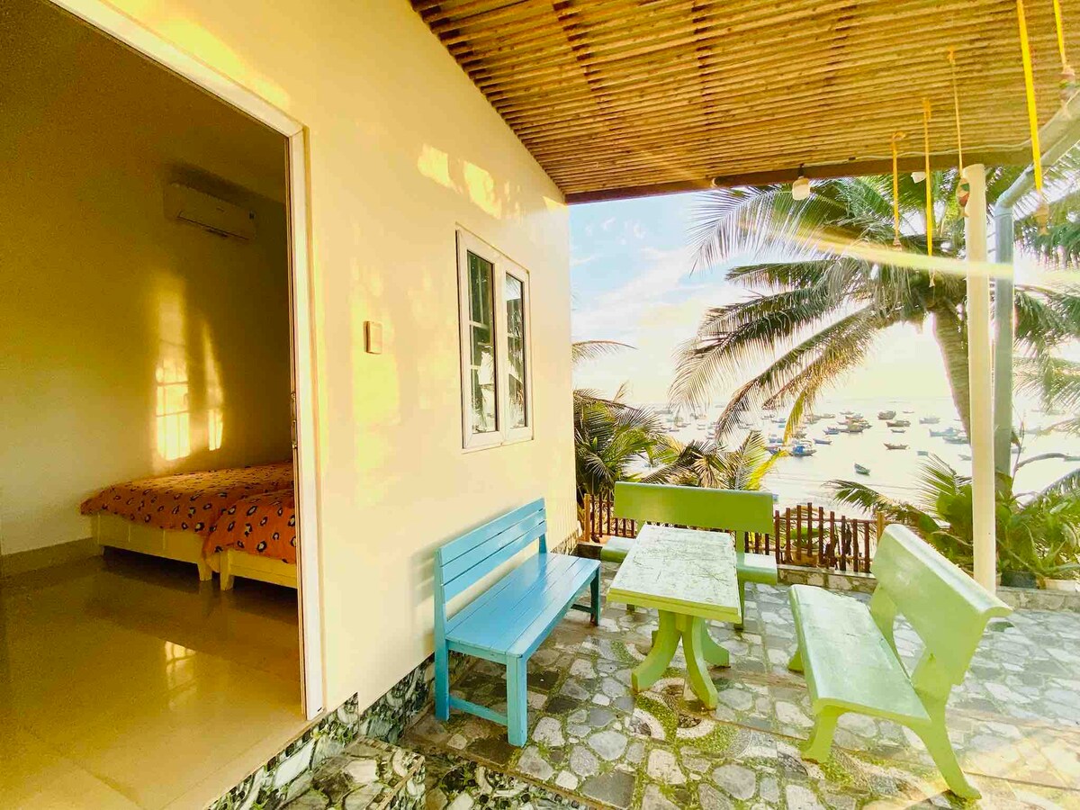 An outdoor seating area features a light green table surrounded by blue and green chairs. The space opens to a view of palm trees and a nearby beach, with boats visible in the distance. Natural light highlights the entrance to a room with a spacious bed.