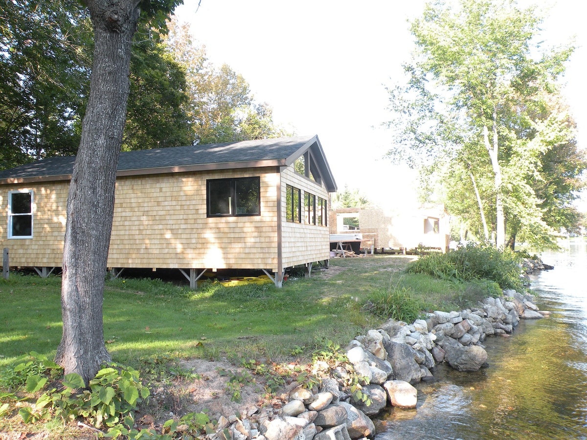 A lakeside cottage is displayed, featuring wooden siding and large windows allowing for natural light. The surrounding greenery and flat lawn lead to a stone border along the water. The tranquil setting is enhanced by the nearby trees and clear lake view.