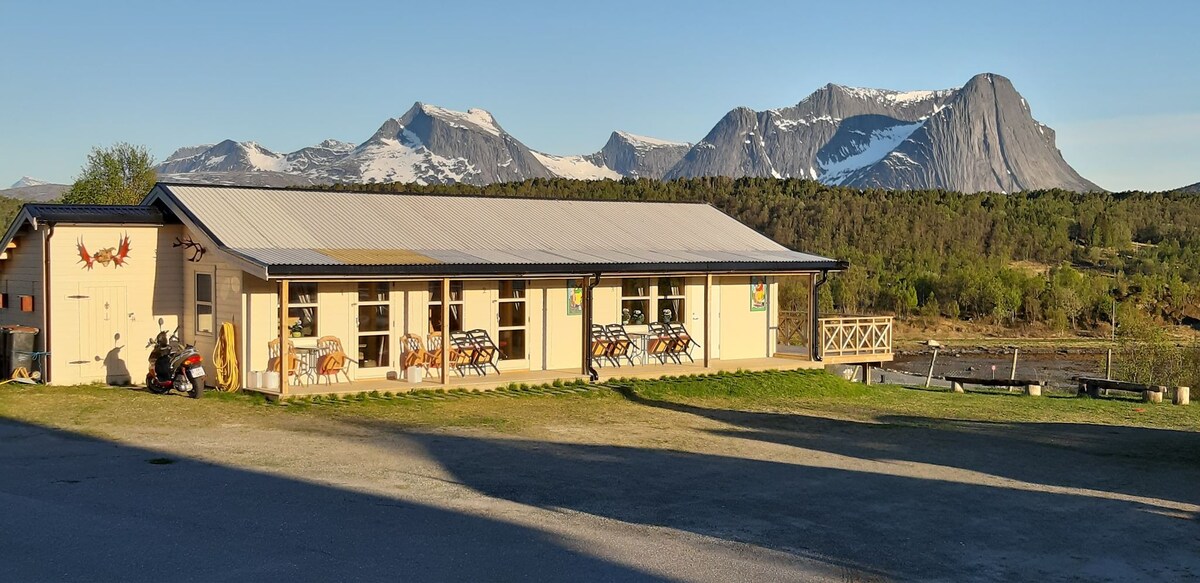 A single-story building is surrounded by lush greenery and mountain peaks in the background. Large windows along the front are complemented by outdoor seating areas. The clear sky highlights the stunning landscape, creating a serene environment.