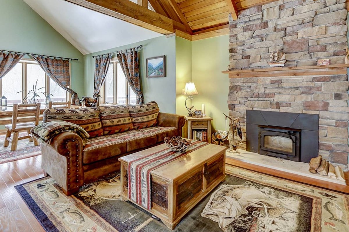 A welcoming living area features a stone-stacked fireplace and handcrafted log furniture. A cozy sofa complements a rustic coffee table, while large windows allow natural light to fill the space. Warm-toned area rugs add texture to the hardwood floor.