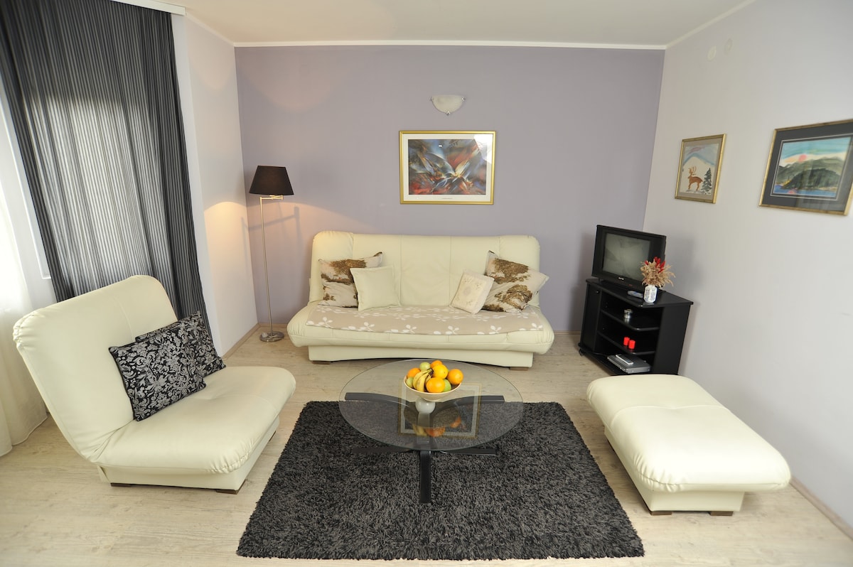 A modern living area is presented with a light-colored sofa and lounge chairs, arranged around a glass coffee table. The room features a textured black rug and artwork on the walls, with natural light filtering in through sheer curtains.
