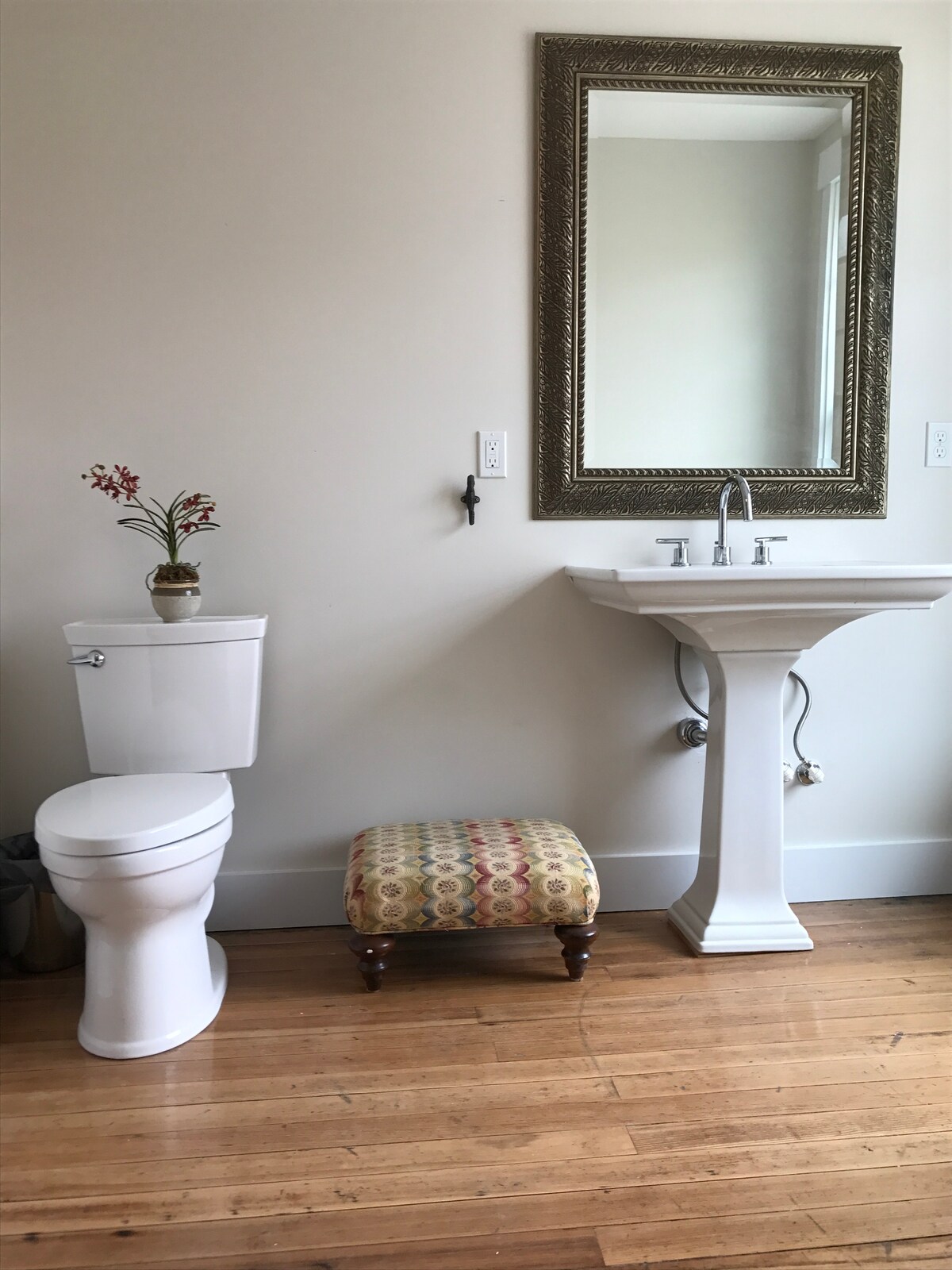 A bathroom is presented with a white pedestal sink beside a mirrored wall. A compact toilet is situated nearby, accented by a decorative stool with a colorful pattern. Natural light brightens the wooden floor, creating a clean and open atmosphere.