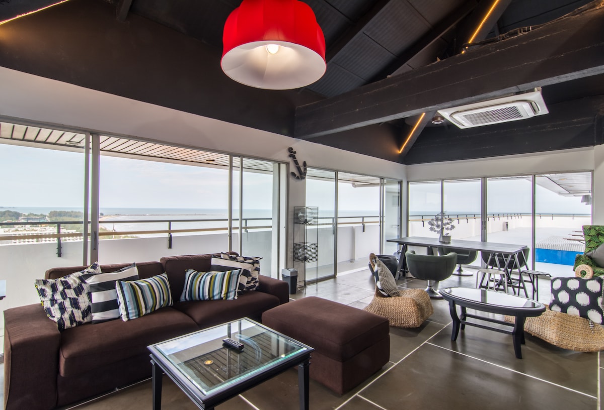 The spacious common area features large windows that offer expansive views of the sea. A comfortable dark sofa with striped cushions is complemented by a glass coffee table. A dining table stands nearby, and natural light fills the space, highlighted by a distinctive red pendant light overhead.