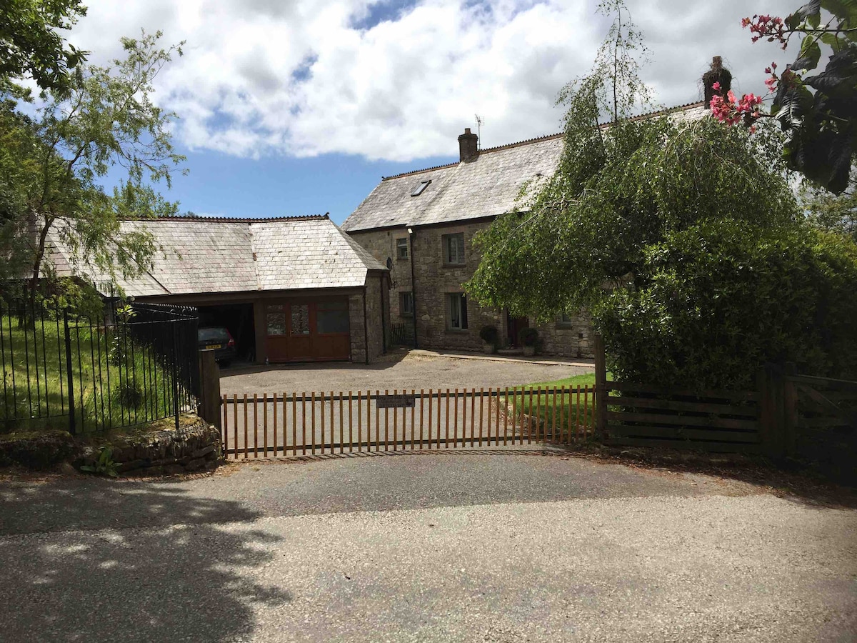 A view of the farm entrance reveals a gated driveway leading to a spacious stone farmhouse with slate roofing. Surrounding greenery and trees create a tranquil environment. The carport is visible on the left, providing covered parking beside the house.