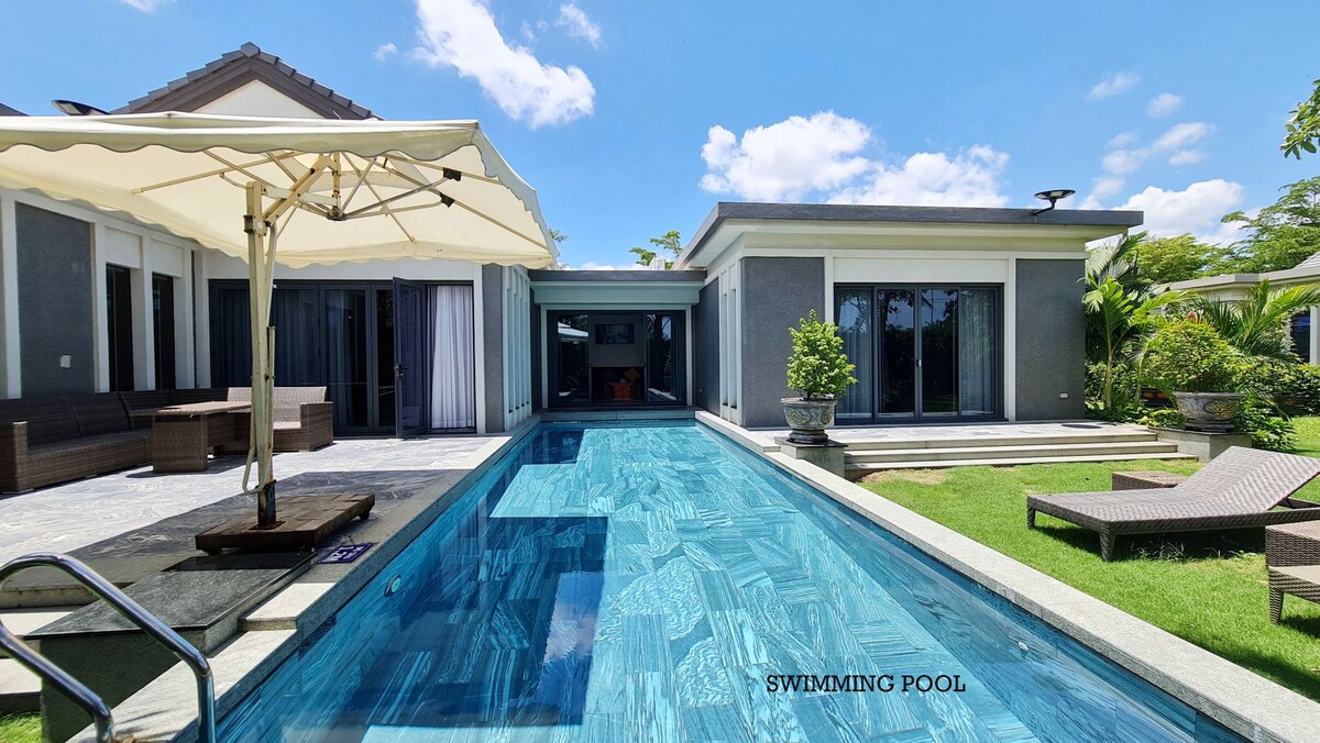 A private swimming pool is showcased, surrounded by elegant outdoor seating, including lounge chairs and a shaded area. The pool’s clear water reflects the blue sky, while the well-maintained landscape features tropical plants, enhancing the tranquil outdoor environment.