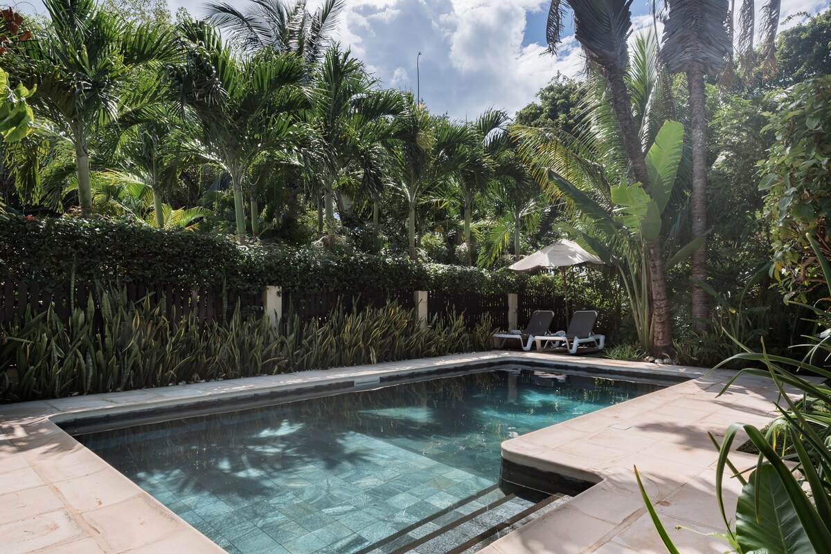 A private pool sits surrounded by lush tropical foliage, offering a serene atmosphere. Sun loungers with an umbrella provide relaxation options near the water, while the tiled deck complements the inviting nature of the pool.