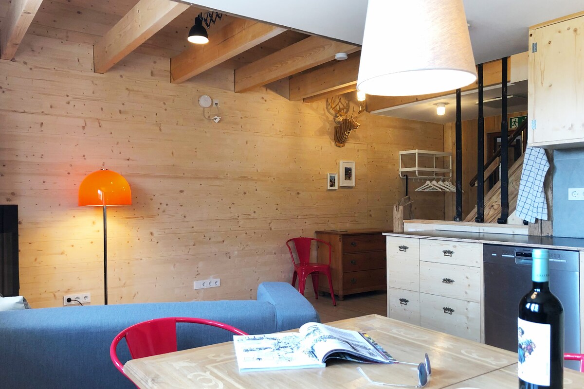 A cozy living space is highlighted by warm wooden walls and exposed beam ceilings. A dining table is set with an open magazine, while a red lamp provides soft lighting. Nearby, a sofa offers a comfortable seating area, with a glimpse of the kitchen area in the background.