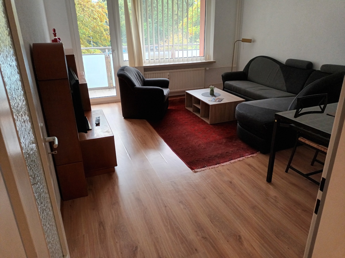 The living area features a comfortable black armchair and a gray sectional sofa, arranged around a wooden coffee table. A red rug adds warmth to the wooden flooring. Large windows allow natural light to illuminate the space, complemented by soft vertical blinds.