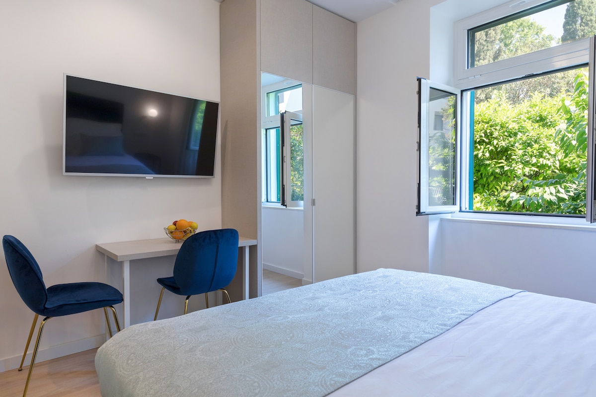A light-filled room features a spacious bed with a soft cover, accompanied by a small table and two upholstered chairs. A flat-screen TV is mounted on the wall, and large windows open to greenery outside, enhancing the bright and airy feel.