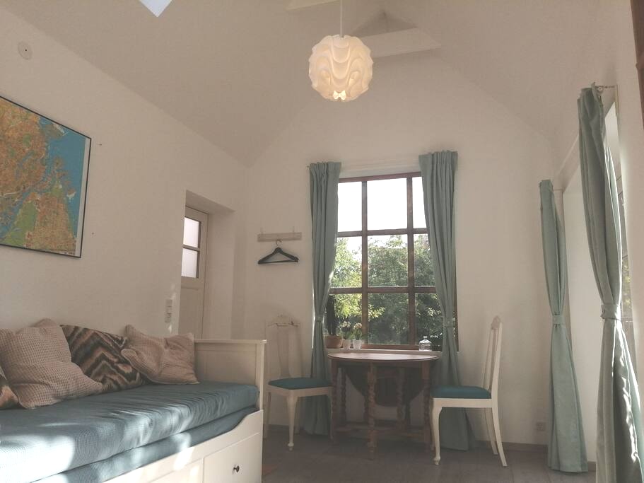 A bright interior features a comfortable seating area with cushions along one side. A small dining table and two chairs are positioned near a window, allowing natural light to illuminate the space. Curtains frame the window, enhancing the airy atmosphere.