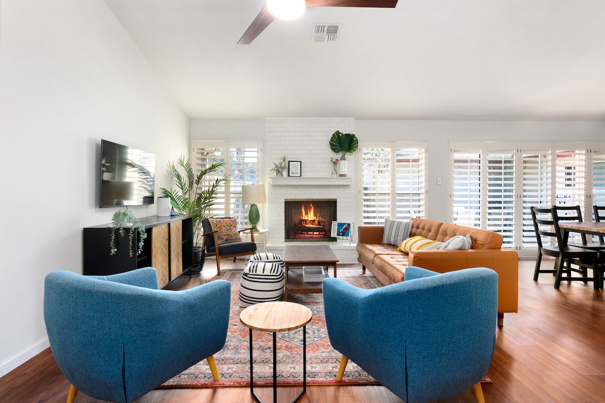 A spacious living room is depicted, featuring a modern fireplace surrounded by light-colored walls. Cozy seating is arranged, including two blue armchairs and a brown sofa. Natural light filters through window shutters, enhancing the warm atmosphere. An area rug adds texture, complemented by a wooden dining set nearby.