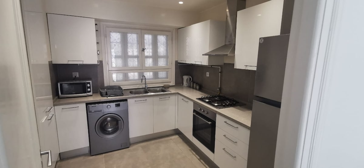 A modern kitchen is equipped with essential appliances, including a microwave, washing machine, and refrigerator. Sleek white cabinets and a contrasting countertop create a clean aesthetic. Natural light filters through the window, illuminating the workspace and enhancing the functional layout.