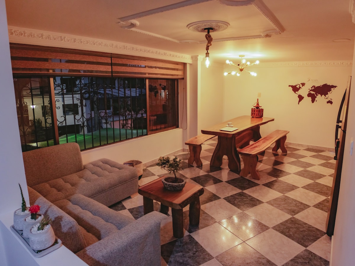A spacious common area is featured, showcasing a large wooden table accompanied by two benches and a coffee table. A gray sofa provides seating, and large windows allow natural light to fill the room, revealing a view of the outdoors. Decorative elements include a small plant on the table and a world map on the wall.
