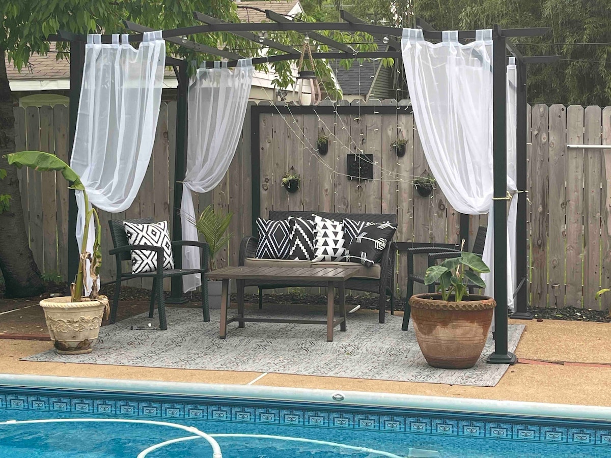 A shaded outdoor seating area is arranged beside a swimming pool. Black and white patterned cushions adorn the chairs and sofas around a central table. Sheer white curtains drape gracefully from a pergola, enhancing the tranquil setting.