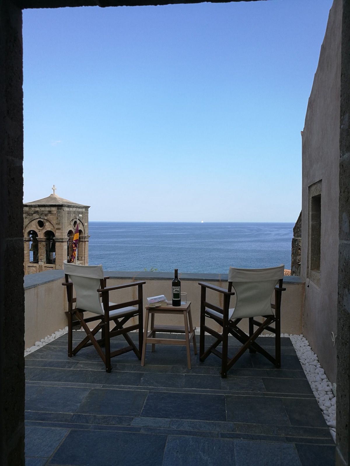 A sea-view terrace is featured, furnished with two wooden chairs and a small table. A bottle of wine and a book are placed on the table. The expansive view of the Aegean Sea is visible, complemented by a clear blue sky.