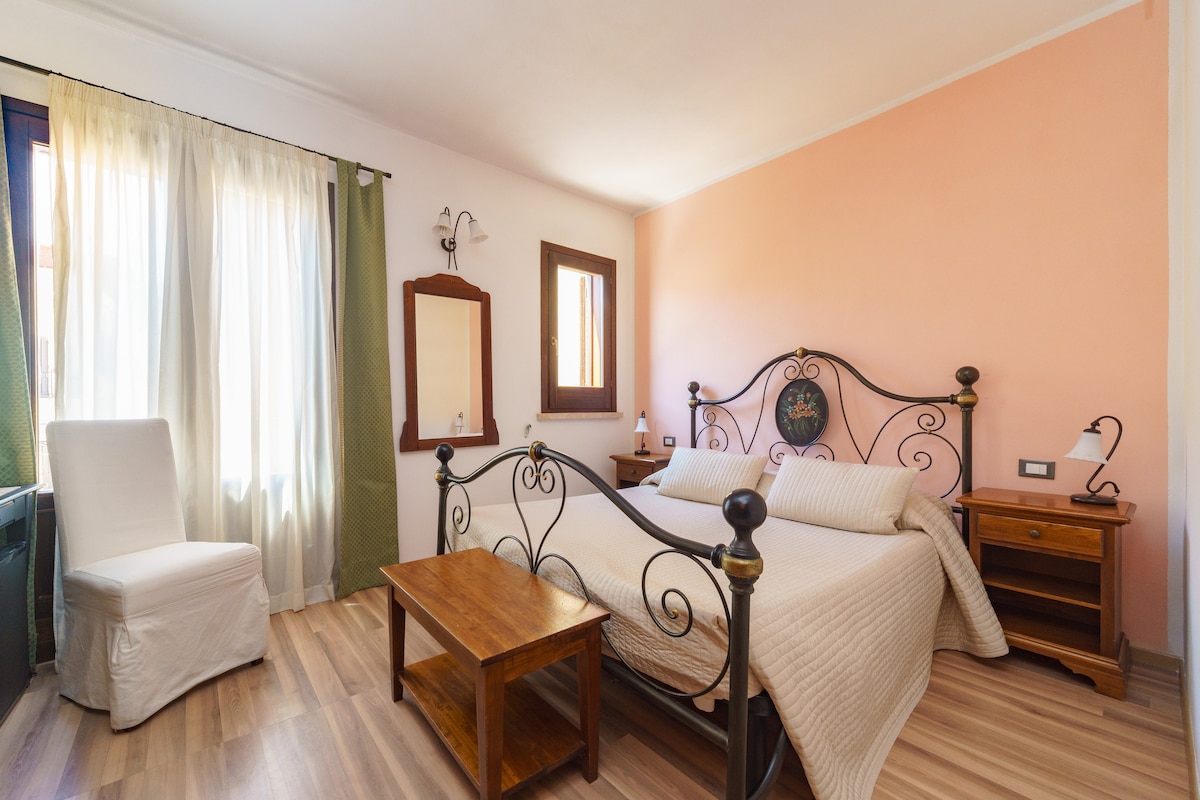A spacious bedroom features a double bed framed by an ornate headboard. Natural light streams through two large windows, complemented by light-colored curtains. A wooden table is situated beside the bed, along with two nightstands that hold lamps, and a full-length mirror reflects the inviting space.