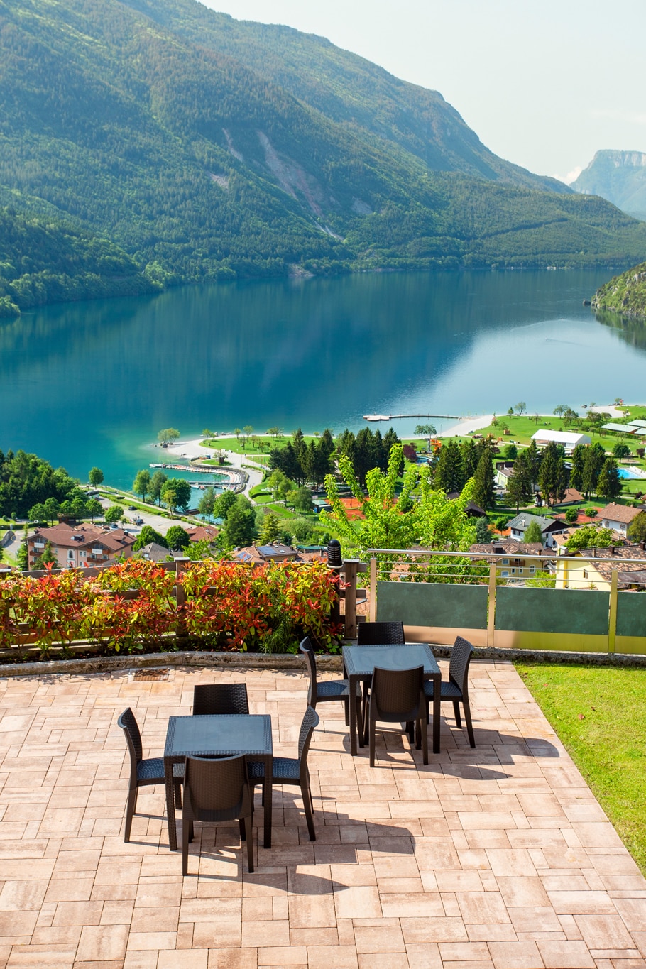 An outdoor terrace features several black tables and chairs, offering a serene view of a clear blue lake surrounded by lush greenery and mountains in the background. The sunlight reflects off the water, enhancing the natural beauty of the landscape.