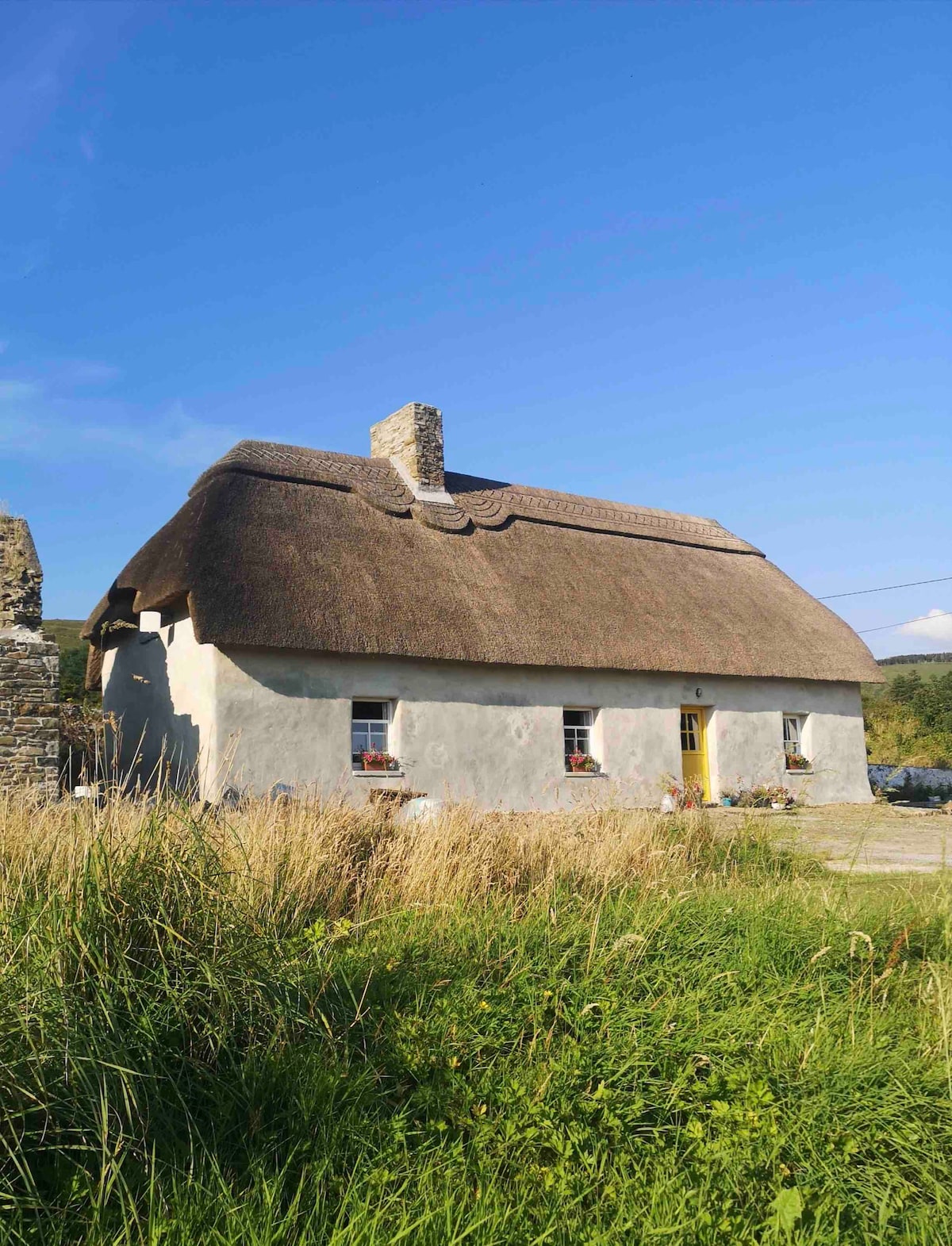 The exterior of a charming thatched cottage is presented, featuring a traditional stone chimney and a bright yellow door. Surrounding green grass and wildflowers enhance the serene rural setting. Clear blue skies stretch overhead, adding to the peaceful atmosphere.