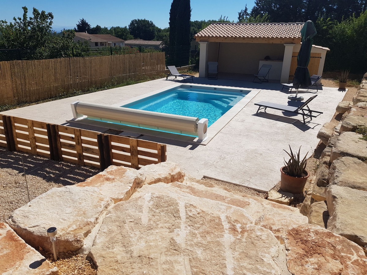 A swimming pool is set within a spacious outdoor area, surrounded by a stone patio. Lounge chairs are positioned nearby, along with a shaded seating area under a roof. Lush greenery and a wooden fence provide privacy, enhancing the inviting vibe of the space.