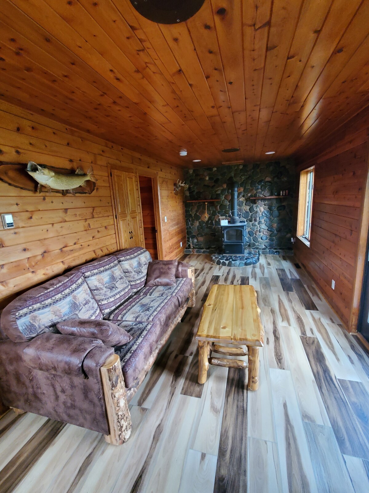 A living area is presented with wooden walls and flooring, featuring a comfortable sofa and a rustic wooden coffee table. A wood stove is positioned near the corner, enhancing warmth. Natural light enters through the window, illuminating the space.