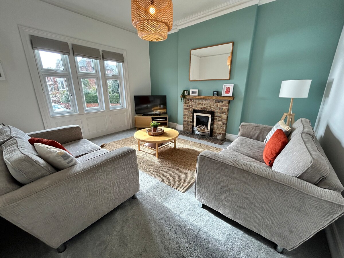 A spacious lounge features two comfortable sofas surrounding a central coffee table. A flat-screen television is positioned on a media unit, with a decorative fireplace and a mirror above it. Natural light enters through large windows adorned with shades.