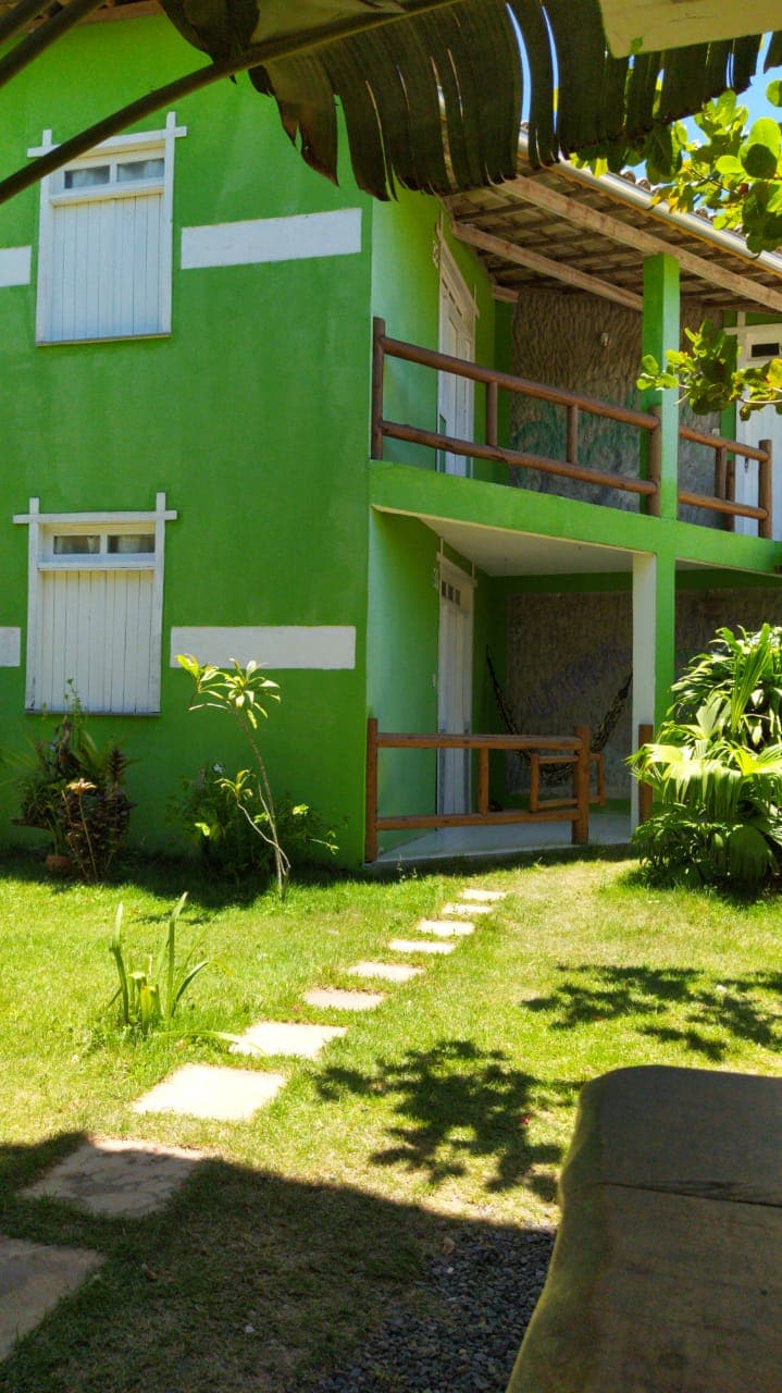 A vibrant green building is surrounded by lush foliage and a neatly trimmed lawn. A pathway of stepping stones leads to the entrance, with wooden balconies featuring railing details visible above. The upper-level windows are framed with white shutters, enhancing the building's charm.