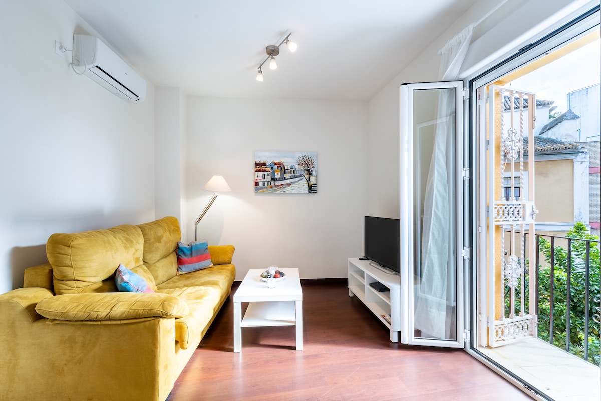 A living area features a comfortable yellow sofa, complemented by a small white coffee table. A television is positioned on a white entertainment unit, while a painting of colorful houses adds character to the space. Natural light enters through the open balcony doors.