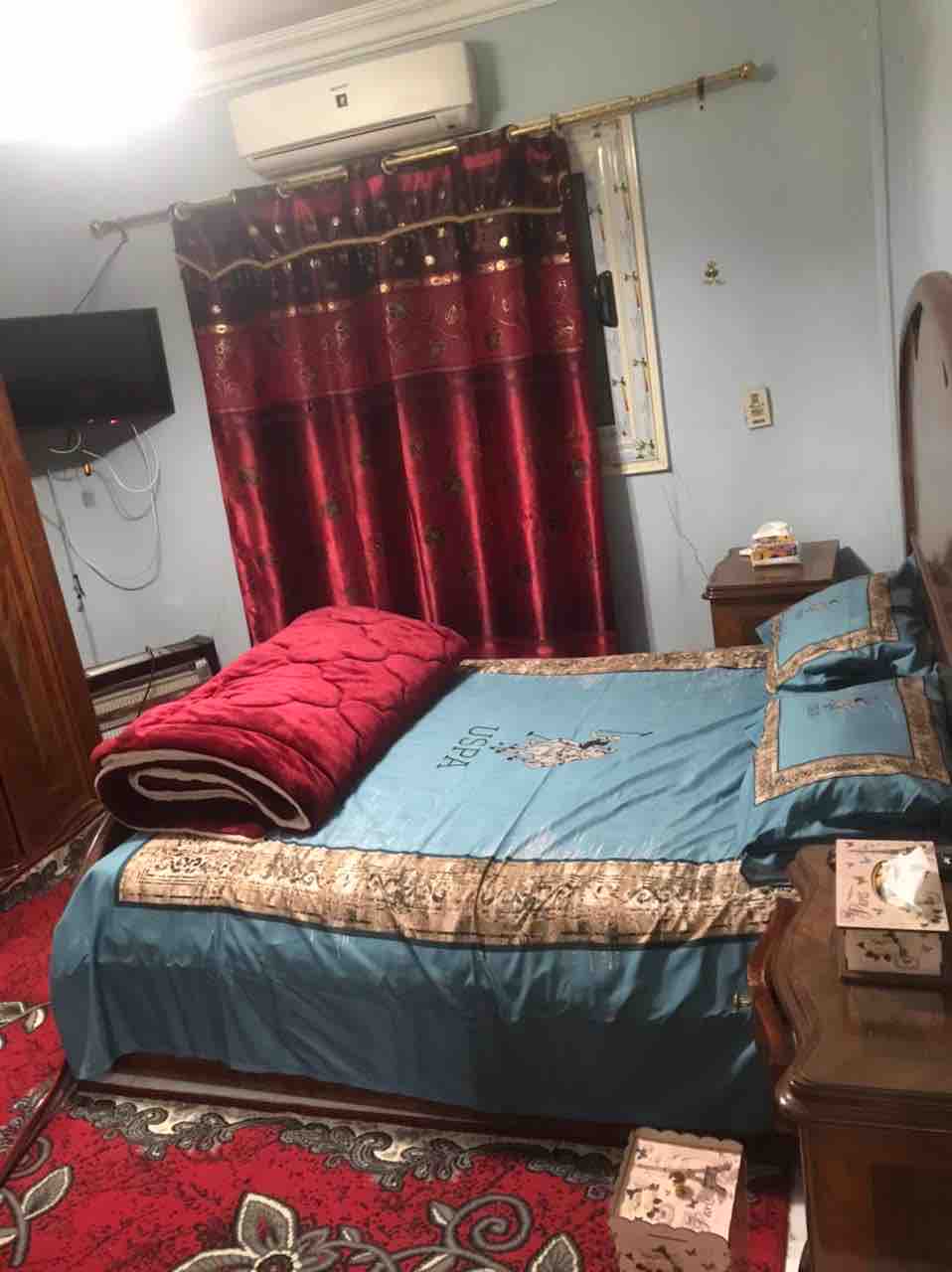 The sleeping area features a spacious bed adorned with vibrant blue and gold bedding. A rich red blanket is neatly placed at the foot of the bed. The room is complemented by a decorative red curtain and a television mounted on the wall, creating a cozy ambiance.