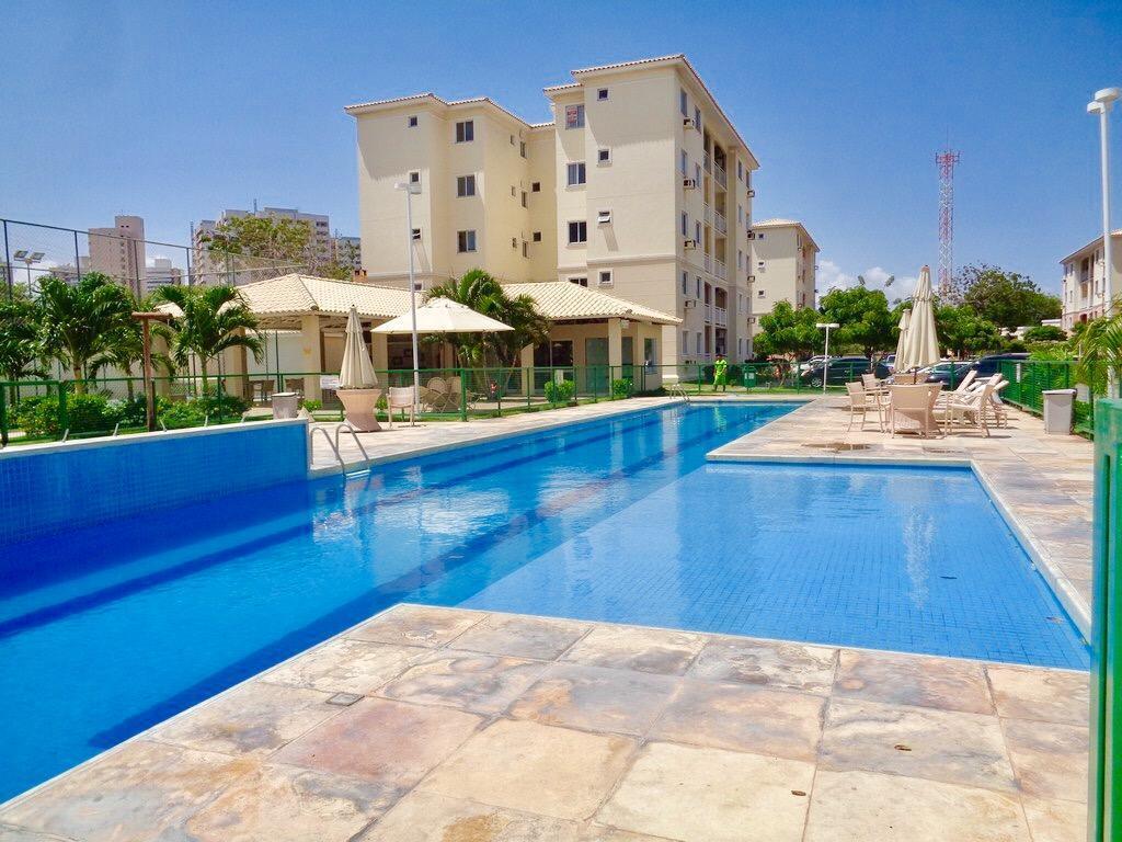 A large swimming pool is surrounded by a tiled patio and well-maintained greenery. Lounge chairs and umbrellas are positioned nearby for relaxation. The building in the background features multiple floors and balconies, contributing to a calming atmosphere within the gated community.
