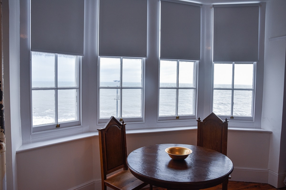 A round wooden dining table is positioned near large bay windows, offering views of the sea. Natural light filters through the white roller blinds, illuminating the space. Two wooden chairs accompany the table, creating a cozy spot for meals or enjoying the scenery.