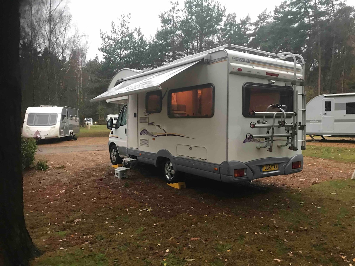 A compact camper van is positioned on a natural site surrounded by trees. The vehicle features an awning extended over the entry, with its back displaying a built-in ladder and bike rack. Nearby, additional campers can be seen in a tranquil outdoor setting.